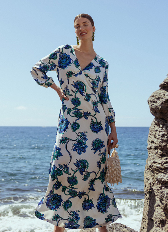 Vrouw in een lange witte jurk met blauwe bloemenprint staat op rotsen aan zee en kijkt ontspannen in de zon. Ze draagt opvallende oorbellen en houdt een kleine tas vast terwijl de golven en heldere lucht de zomerse sfeer versterken.