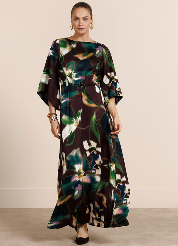 Vrouw draagt een lange jurk met kleurrijke print en lange mouwen, gecombineerd met sandalen. De outfit laat een speelse en elegante stijl zien die geschikt is voor een zomerse setting.