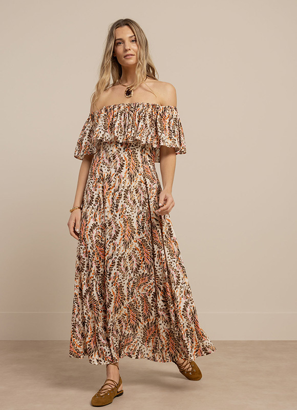 Vrouw staat tegen een neutrale wand en draagt een bruine jumpsuit met korte mouwen en een ceintuur in de taille. De outfit geeft een verfijnde en minimalistische uitstraling met een zachte kleur.