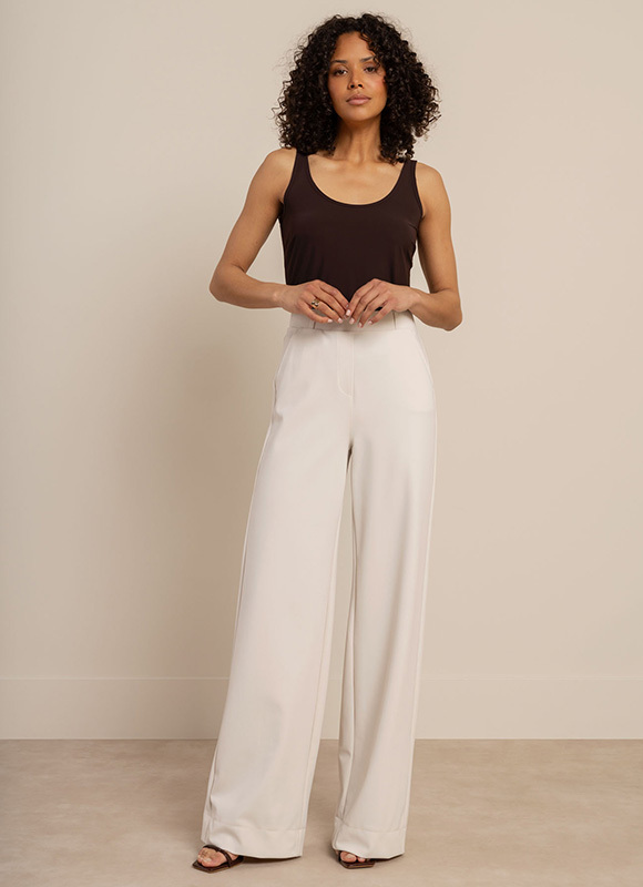 Vrouw staat buiten onder bomen en draagt een beige mouwloze jumpsuit met strikceintuur en sandalen. De outfit laat zien hoe een lichte jumpsuit zorgt voor een elegante en zomerse look.