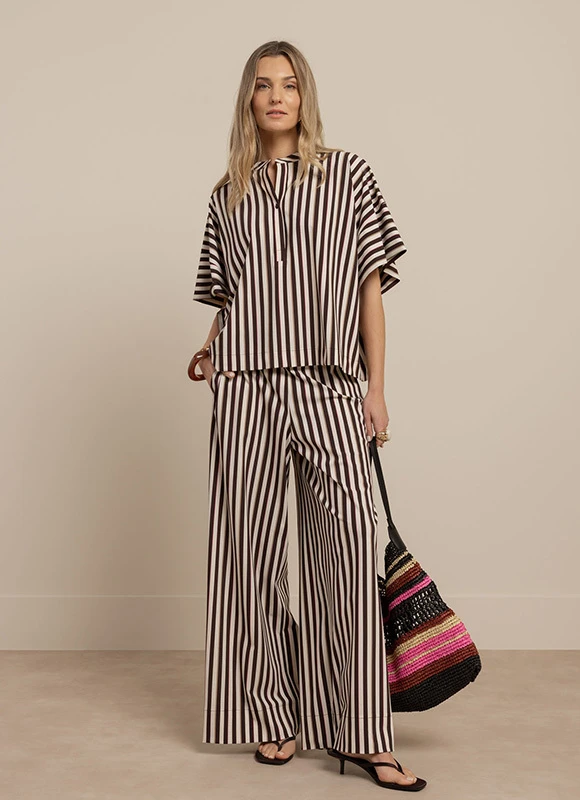 Vrouw draagt een bruin krijtstreep pak met een ceintuur en staat rechtop tegen een neutrale achtergrond. De klassieke strepen en getailleerde pasvorm geven de outfit een krachtige en elegante uitstraling.