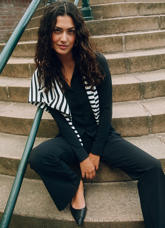 Vrouw in een losse zwarte jurk met knopen staat tegen een neutrale achtergrond en kijkt zelfverzekerd naar voren terwijl ze haar hand bij haar gezicht houdt. De minimalistische styling en comfortabele pasvorm geven de outfit een tijdloze en moeiteloze uitstraling.