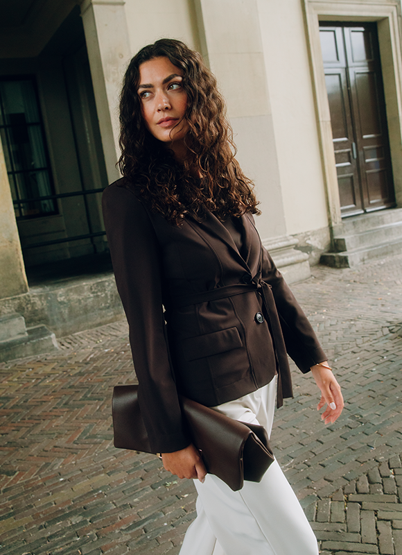 Twee vrouwen dragen outfits in donkerbruine tinten en staan zelfverzekerd tegen een neutrale achtergrond. De combinatie van een gestreepte top met short en een elegante set met riem zorgt voor een moderne en stijlvolle uitstraling.