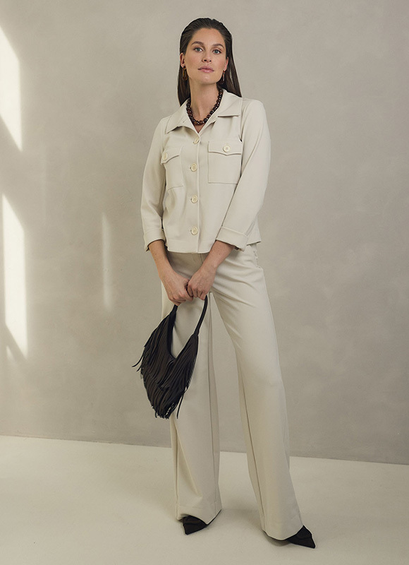 Vrouw draagt een lichtblazer over een donkere outfit en staat rechtop tegen een neutrale achtergrond. De combinatie van lichte en donkere tinten zorgt voor een elegante en moderne uitstraling.