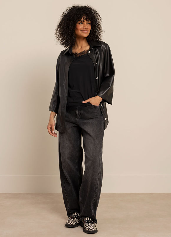 Vrouw staat voor een klassiek gebouw en draagt een donker faux leather jasje gecombineerd met een blouse met print en een wijde geruite broek. De combinatie benadrukt hoe een faux leather item eenvoudig kan worden gestyled als statement piece binnen een outfit.