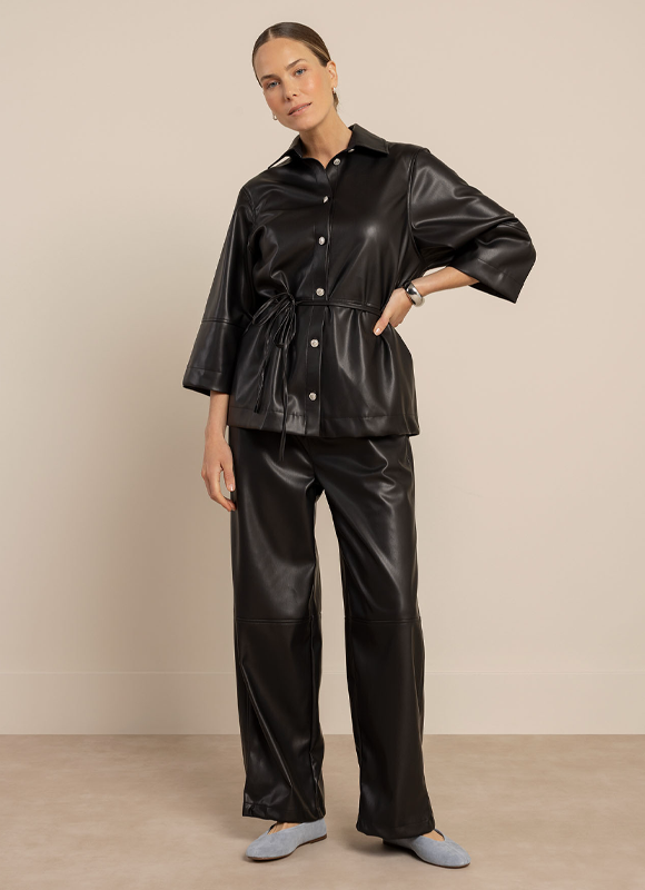 Vrouw loopt over een met bomen omzoomd pad en draagt een roze blouse met pofmouwen en een donkere faux leather broek met enkellaarsjes en handtas. De outfit laat zien hoe faux leather een stijlvolle en draagbare keuze is voor een elegante dagelijkse look.