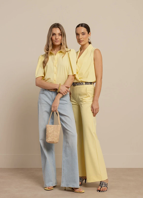Twee vrouwen staan naast elkaar tegen een neutrale achtergrond en dragen beide een outfit in zachte gele tinten. Eén vrouw combineert een gele blouse met een lichte jeans en een geweven tas terwijl de ander een mouwloze gele jumpsuit draagt met een riem en sandalen met zebraprint.