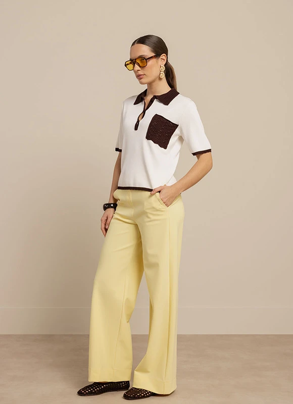 Vrouw draagt een witte polo met donkerbruine kraag en borstzak gecombineerd met lichtgele wijde broek en bruine accessoires terwijl ze een zonnebril draagt. De outfit laat zien hoe geel gecombineerd kan worden met warme bruintinten voor een stijlvolle en moderne look.