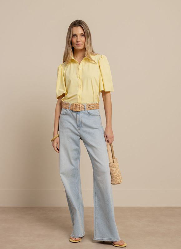 Vrouw draagt een lichtgele blouse met korte mouwen gecombineerd met een lichte jeans een gevlochten riem en een kleine geweven tas. De zachte kleuren geven de outfit een frisse en zomerse uitstraling.