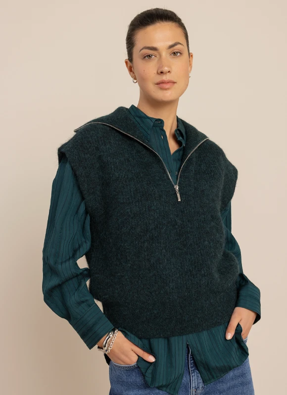 Vrouw draagt een beige broek met een witte blouse, een vest en een losgeknoopte stropdas, gecombineerd met hakken. De outfit benadrukt een speelse en verfijnde mix van klassiek en casual styling.