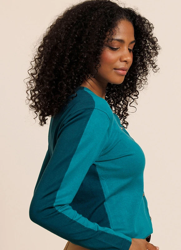 Vrouw zit ontspannen op een bank en draagt een taupekleurige outfit met een lichtgrijze sjaal nonchalant over haar schouders. De zachte materialen en neutrale tinten geven de look een comfortabele en stijlvolle uitstraling.
