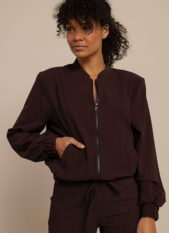 Vrouw draagt een donkerbruine set met een ritsjack en broek en staat ontspannen met haar hand in haar zak tegen een neutrale achtergrond. De minimalistische styling en diepe kleur geven de outfit een moderne en sportieve uitstraling.