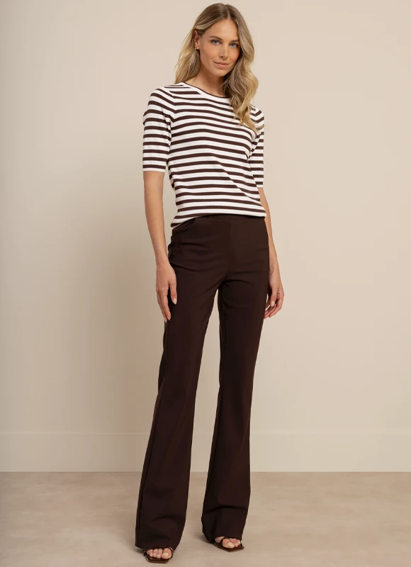 Vrouw draagt een witte top met een beige flared broek en staat met haar rug naar de camera terwijl ze een tas vasthoudt. De pasvorm van de broek en de lengte worden benadrukt door de elegante en gestrekte houding.