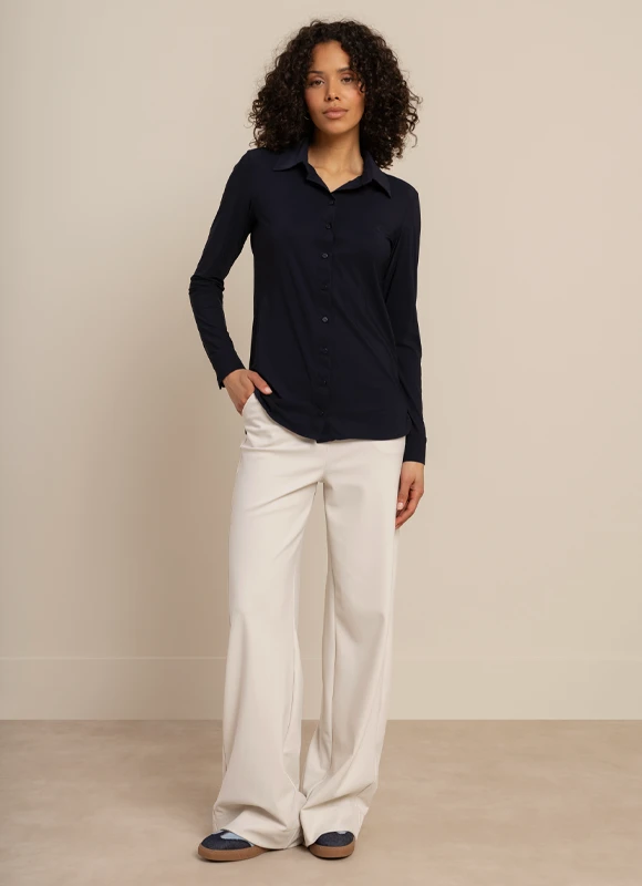 Vrouw draagt een donkerblauwe outfit met een wijde broek en staat met haar rug naar de camera terwijl ze een tas in haar hand houdt. De soepele stof en lange lijnen geven de look een elegante en moderne uitstraling.