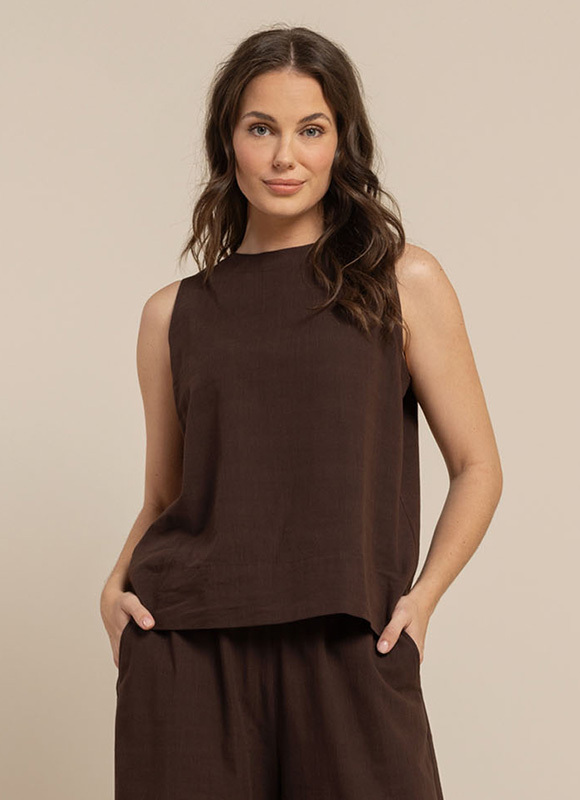 Vrouw staat buiten in een zonnige setting en draagt een luchtige outfit met een beige top, een wit opengewerkt blouse en een gestreepte broek. De combinatie laat een ontspannen en stijlvolle zomerlook zien met natuurlijke tinten.