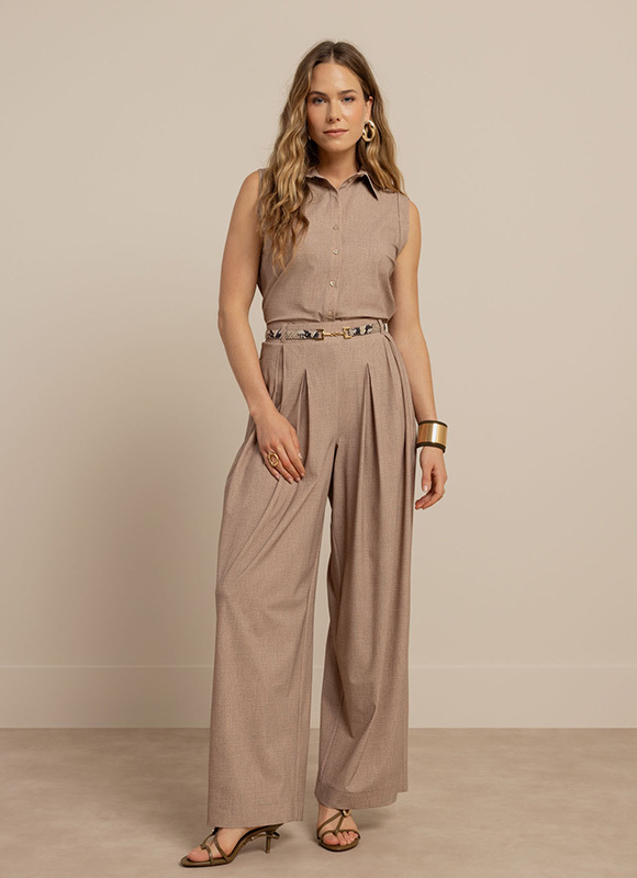 Vrouw staat tegen een witte wand en draagt een beige gestreepte set met een losvallende blouse en wijde broek. De outfit benadrukt een comfortabele en elegante co ord look met een rustige uitstraling.