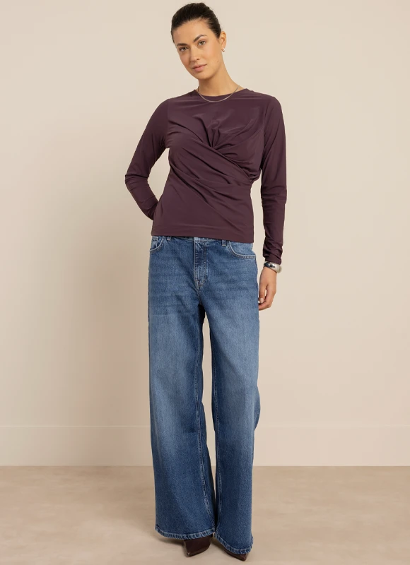 Vrouw draagt een zachte bruine trui over een kleurrijke blouse en glimlacht terwijl ze haar arm omhoog houdt. De mix van texturen en lagen zorgt voor een warme en speelse uitstraling.
