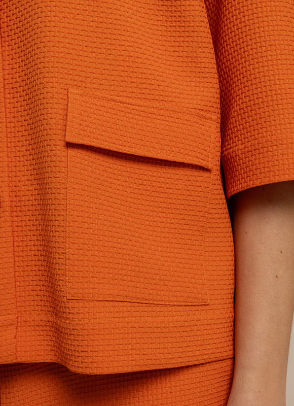Close up van een oranje structuurstof top met korte mouwen en een grote opgestikte zak aan de voorkant. De foto benadrukt de textuur en warme kleur van het kledingstuk.