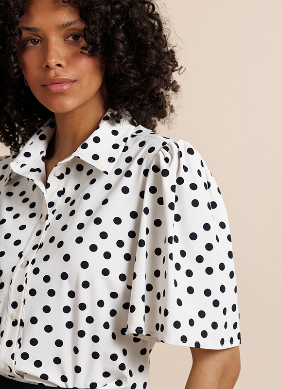 Vrouw draagt een blouse met print en een donkere broek en staat zelfverzekerd in een klassieke interieursetting. De combinatie van het patroon en de elegante houding geeft de outfit een krachtige en modieuze uitstraling.