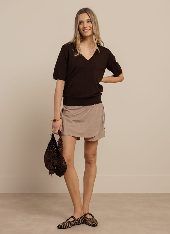 Vrouw draagt een donkerbruine top met korte mouwen en een beige asymmetrische rok gecombineerd met gevlochten ballerina’s en een donkere gehaakte tas. De outfit heeft een zachte en natuurlijke uitstraling met warme neutrale tinten.