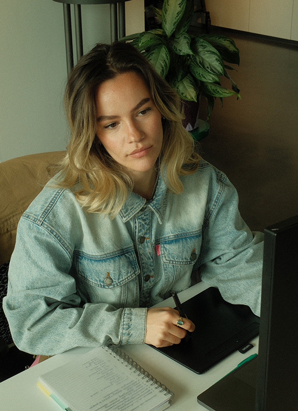 Vrouw zit aan een bureau en werkt geconcentreerd op een tekentablet terwijl ze een pen vasthoudt en naar het scherm kijkt. Haar denim jas en de notities op tafel benadrukken een creatieve werkomgeving met focus op ontwerpen of digitale schetsen.