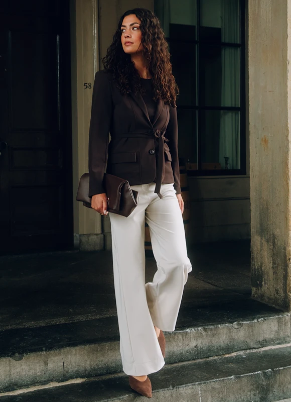 Vrouw staat tegen een warme bruine achtergrond en draagt een donkerbruine jumpsuit met wijde pijpen en hakken, gecombineerd met een tas. De look laat zien hoe een jumpsuit een elegante en complete outfit vormt met minimale styling.