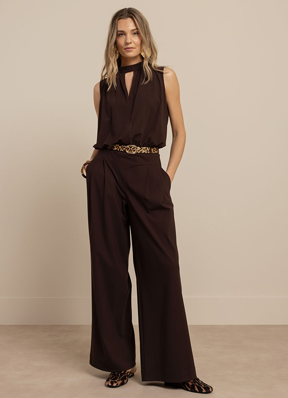 Vrouw draagt een donkerbruine mouwloze jumpsuit met wijde pijpen en een subtiele keyhole halslijn. Een smalle dierenprint riem en platte schoenen geven de elegante outfit een speelse touch.