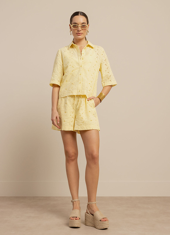 Vrouw draagt een lichtgele co-ord set met korte blouse en bijpassende short met subtiele print. De outfit is gecombineerd met plateau sandalen, gouden sieraden en zonnebril voor een frisse zomerse look.