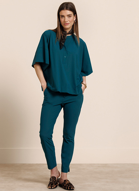 Vrouw leunt tegen een stenen muur en draagt een zwart witte blouse met botanische print en een zwarte short, gecombineerd met sandalen. De luchtige outfit laat zien hoe een print blouse stijlvol werkt in een zomerse setting.