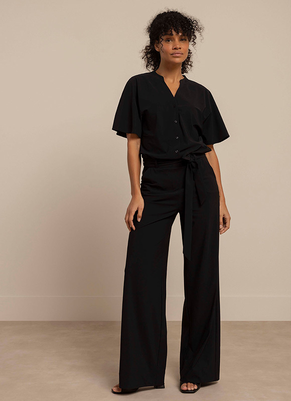 Drie vrouwen dragen zwarte outfits variërend van blazer tot broek en sneakers, staand in een moderne architectonische setting. De looks tonen hoe zwart veelzijdig te combineren is van casual tot zakelijk.