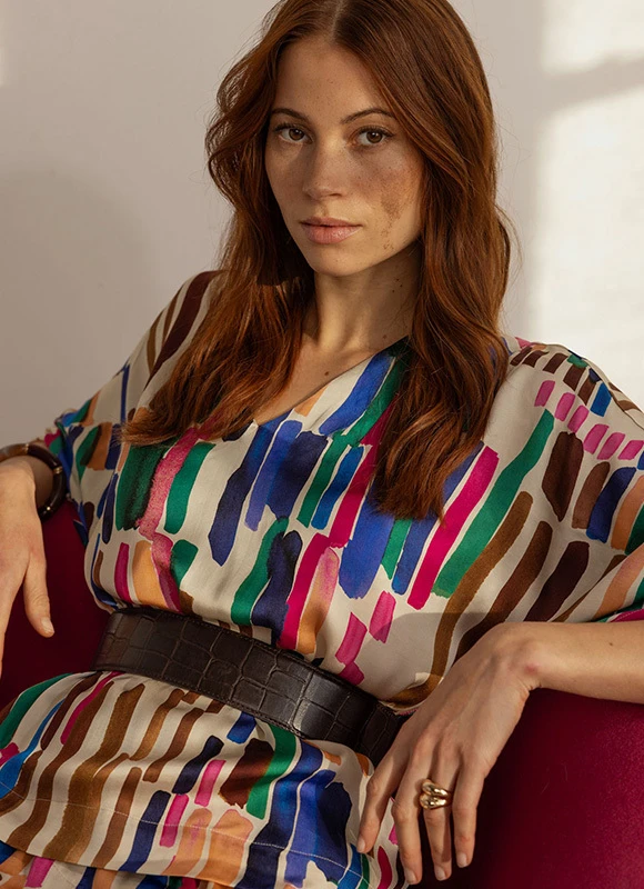 Vrouw zit ontspannen in een stoel en draagt een kleurrijke jurk met abstracte strepen in verschillende tinten. De combinatie van het patroon en de rustige houding benadrukt de speelse en expressieve kracht van kleur.