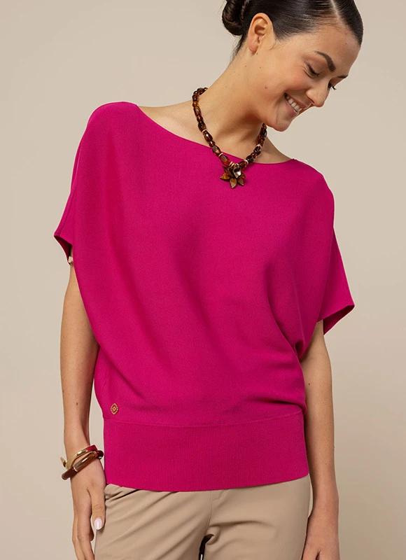 Vrouw draagt een felroze top en kijkt glimlachend naar beneden terwijl ze haar hoofd licht kantelt. De levendige kleur en eenvoudige styling leggen de nadruk op de frisse en energieke uitstraling van de outfit.