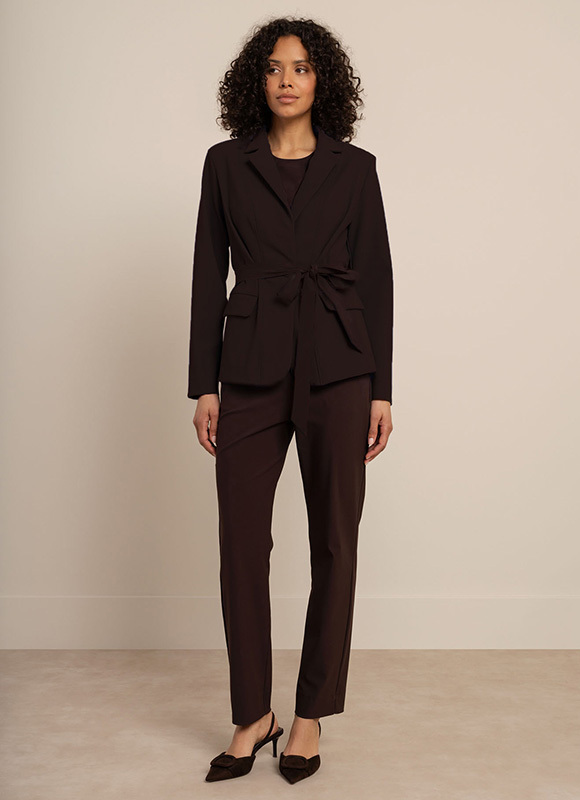 Vrouw draagt een donkerbruine jumpsuit met wijde pijpen en hakken en staat tegen een neutrale achtergrond. De outfit toont een elegante en comfortabele keuze voor zowel werk als vrije tijd.