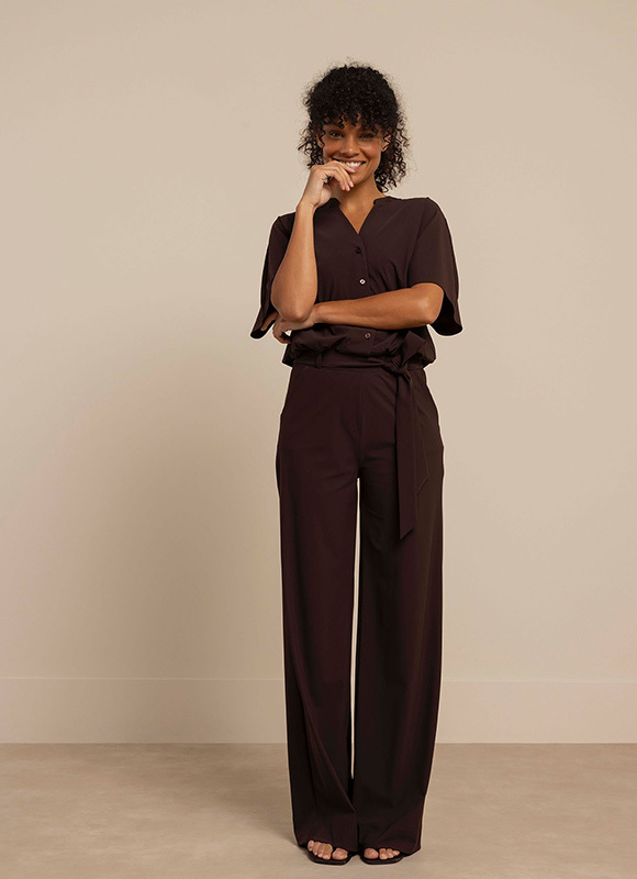 Vrouw staat rechtop en draagt een donkerbruine jumpsuit met korte mouwen en wijde pijpen. De ton-sur-ton look geeft een elegante en moderne uitstraling.