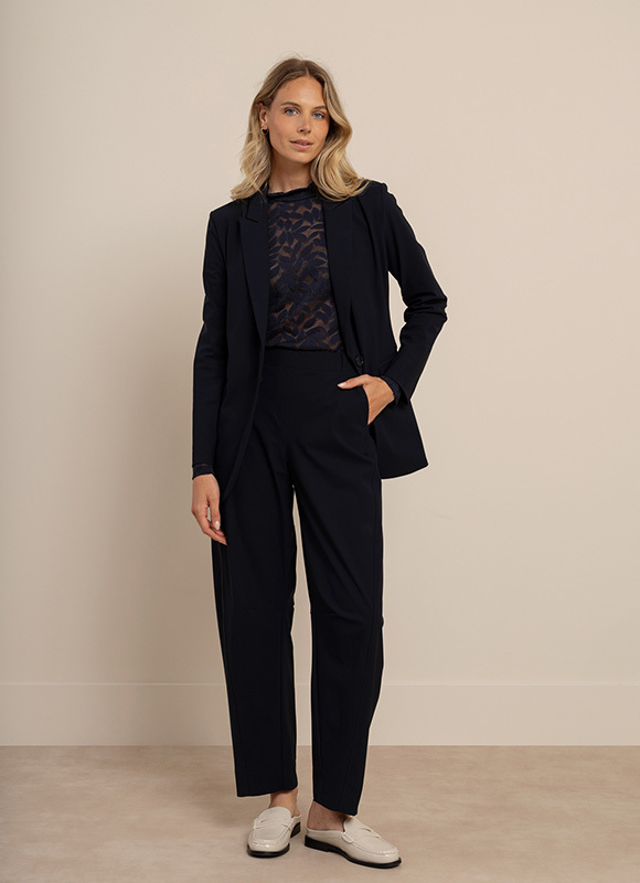 Vrouw draagt een donkerblauw pak met een wit T-shirt en sneakers en loopt ontspannen naar voren. De look benadrukt hoe een bomber jacket een sportieve twist geeft aan een klassieke outfit.