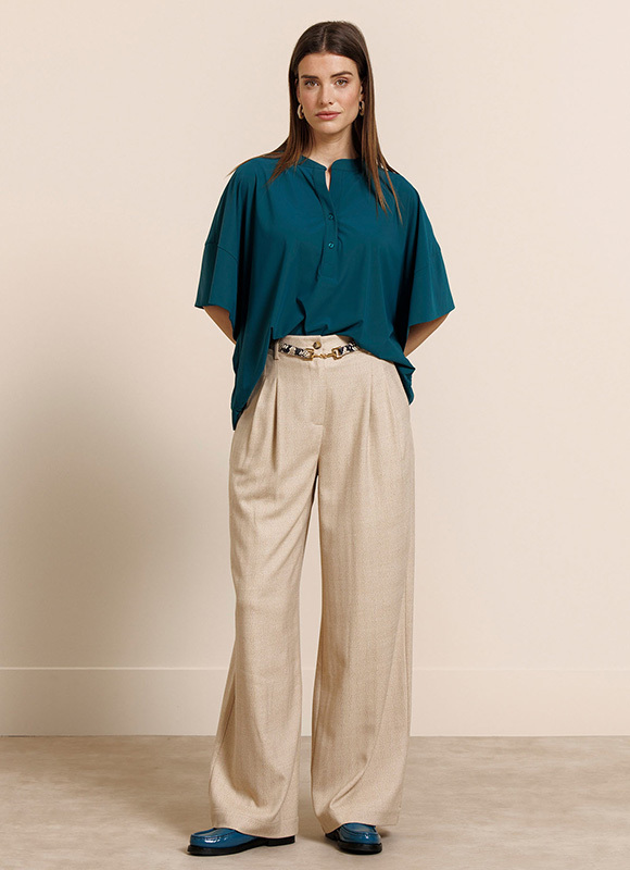 Vrouw draagt een beige blouse met een limegroene broek en houdt een tas vast terwijl ze rechtop staat tegen een neutrale achtergrond. Het contrast tussen de zachte en felle tinten geeft de outfit een frisse en moderne uitstraling.