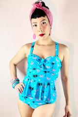 Front View of Pin-up Model Wearing a pink scarf  and the Retro Vintage inspired Midge Swimsuit set with a sky blue peplum tankini that features molded cups and High waist bikini bottom both with  pink flamingo and black palm tree tiny prints.