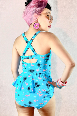 Back View of Pin-up Model Wearing the Retro Vintage inspired Midge Swimsuit set with a sky blue peplum tankini that features molded cups and High waist bikini bottom both with  pink flamingo and black palm tree tiny prints, showing cross back.