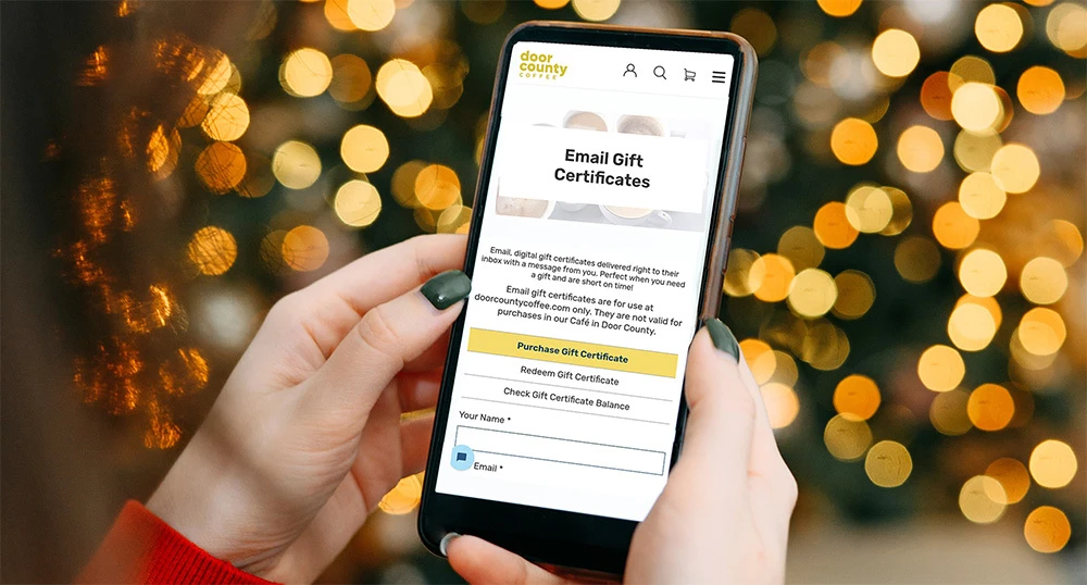 a festive scene containing wrapped gifts, a modern computer keyboard, and a spartphone opened to email gift certificates webpage