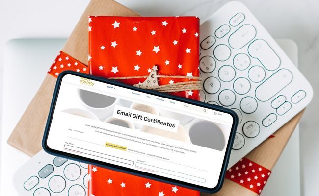 a festive scene containing wrapped gifts, a modern computer keyboard, and a spartphone opened to email gift certificates webpage