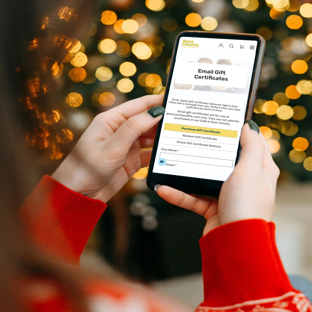 a festive scene containing wrapped gifts, a modern computer keyboard, and a spartphone opened to email gift certificates webpage