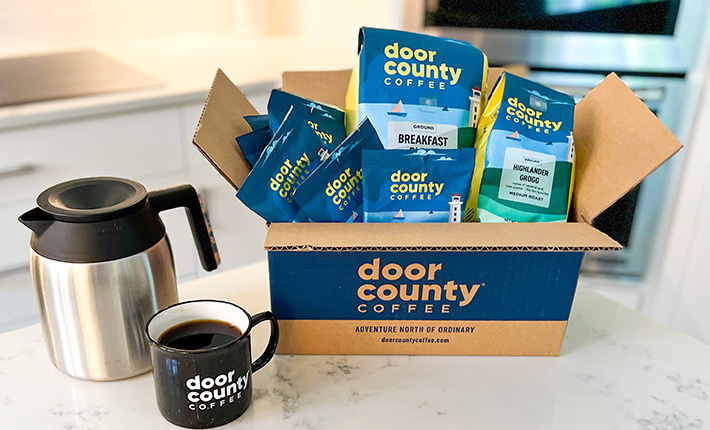 a door county coffe box loaded with coffee products beside a filled coffee mug on a countertop