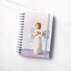 Front view: spiral-bound notebook featuring front of figure with wire wings in cream dress carved with natural symbols, hands crossed under gold heart on chest