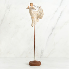 Far shot of angel figurine dressed in white dress dancing with arms and legs out - she is on a brown wooden stand