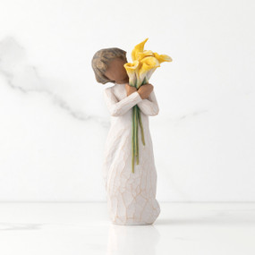 Front view of standing figure in cream dress with darker skin holding yellow calla lilies in her arms