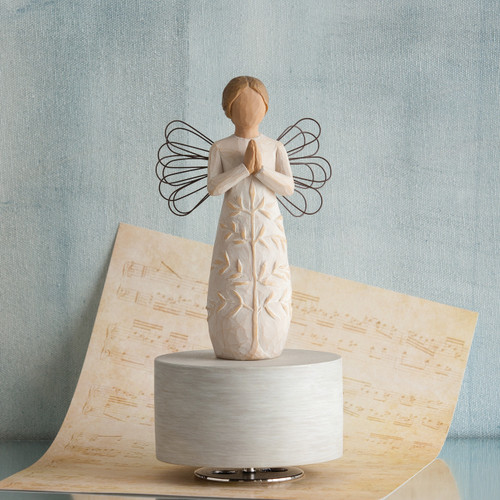 Angel figurine in white dress with hands folded in prayer - she has black wire wings and is standing on round white plaque - background is light blue with a tan colored music sheet below her