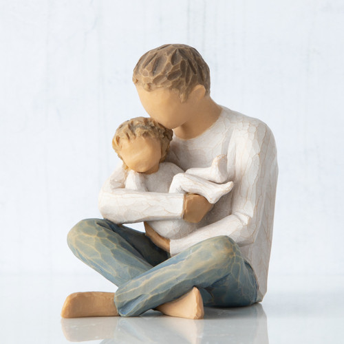 Front view, seated male figure in cream shirt and blue jeans, holding young child in his arms; child in cream onesie