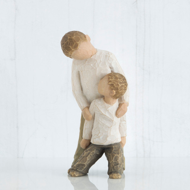 Two small boy figurines wearing white shirts and brown pants -one is standing over holding the other