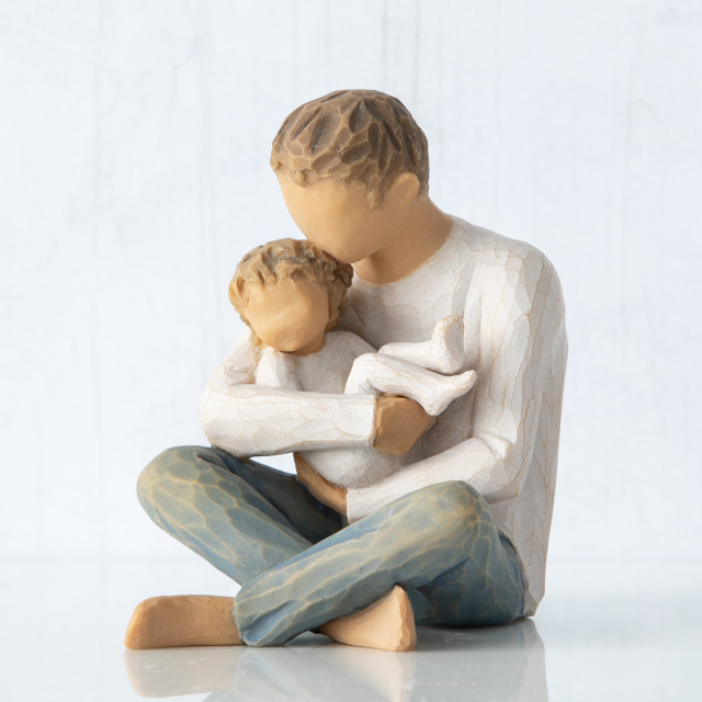 Front view, seated male figure in cream shirt and blue jeans, holding young child in his arms; child in cream onesie