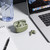Green wireless earbuds with charging case on desk, one earbud outside, beside laptop.